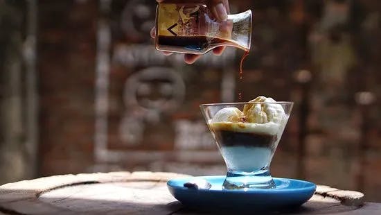 A hand pours espresso over a glass of vanilla ice cream in a blue saucer, making an affogato dessert. The background is blurred, highlighting the wooden table and the contrast between the glass and the dark liquid.