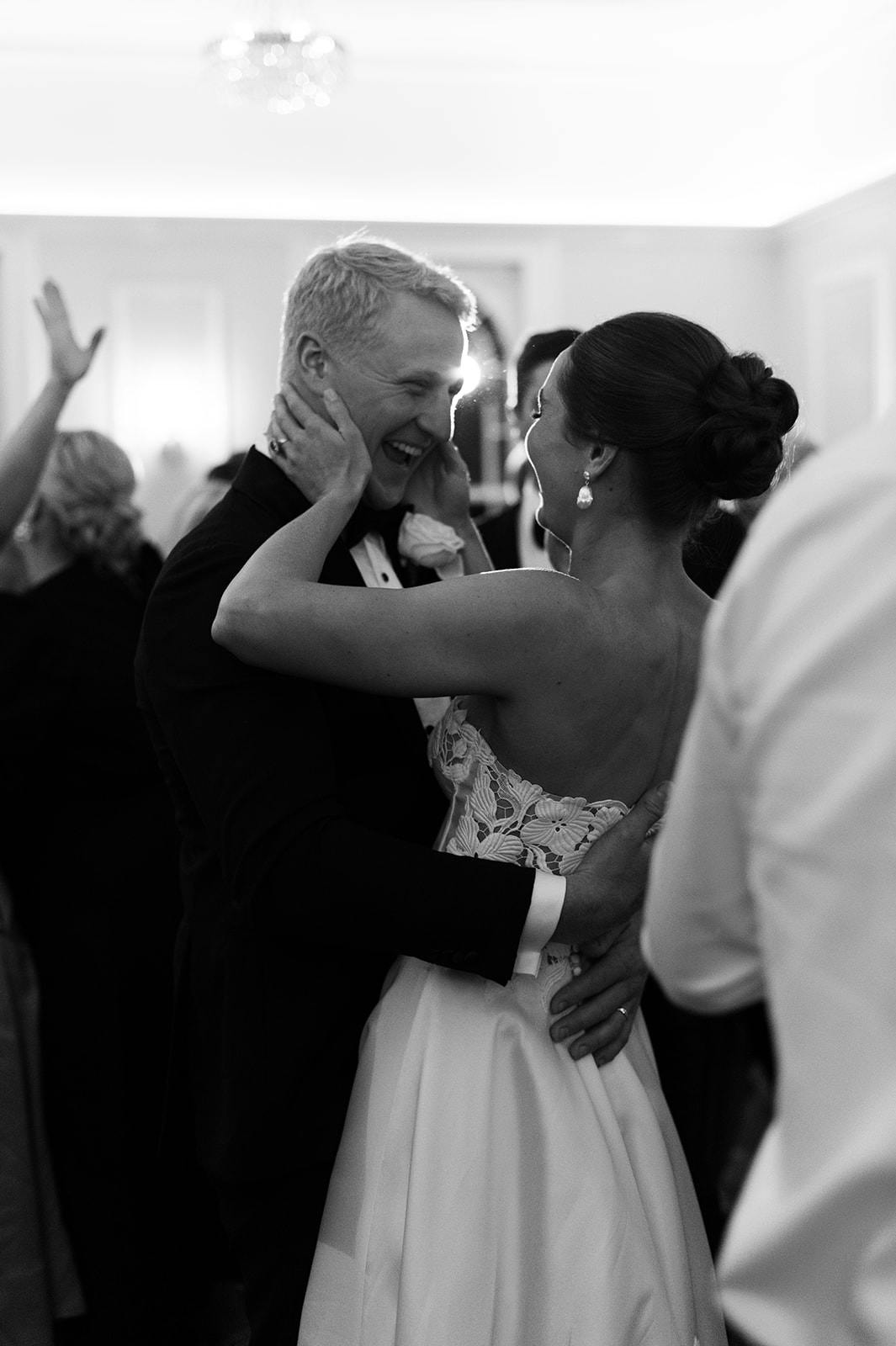 A bride and groom share a joyous moment, laughing and holding each other closely on a dance floor. The bride, wearing an elegant strapless dress, and the groom, in a suit, are surrounded by other guests. The scene is captured in black and white.