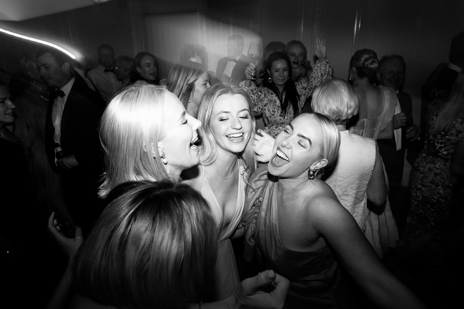 A black and white photo of a lively, crowded party. Three women, smiling and with eyes closed, are in the foreground. They appear to be enjoying themselves, surrounded by a joyful crowd in the background. One woman holds a drink while others dance and celebrate.
