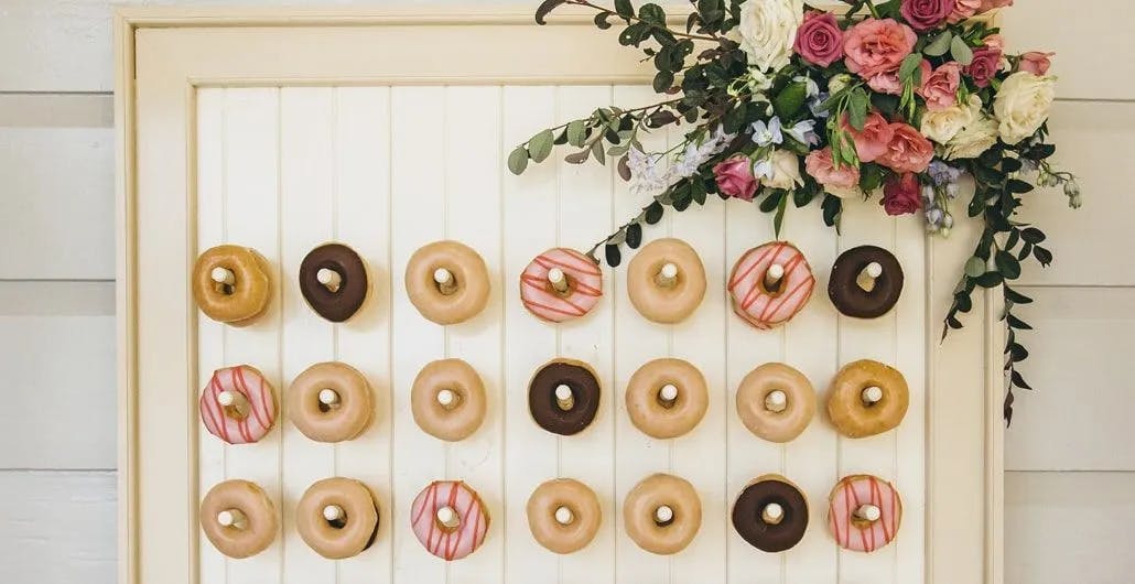 A decorative board with rows of assorted donuts neatly placed on pegs, including glazed, chocolate, and pink-striped varieties. The board is embellished with a floral arrangement of pink, white, and purple flowers, along with green foliage on the top right corner.