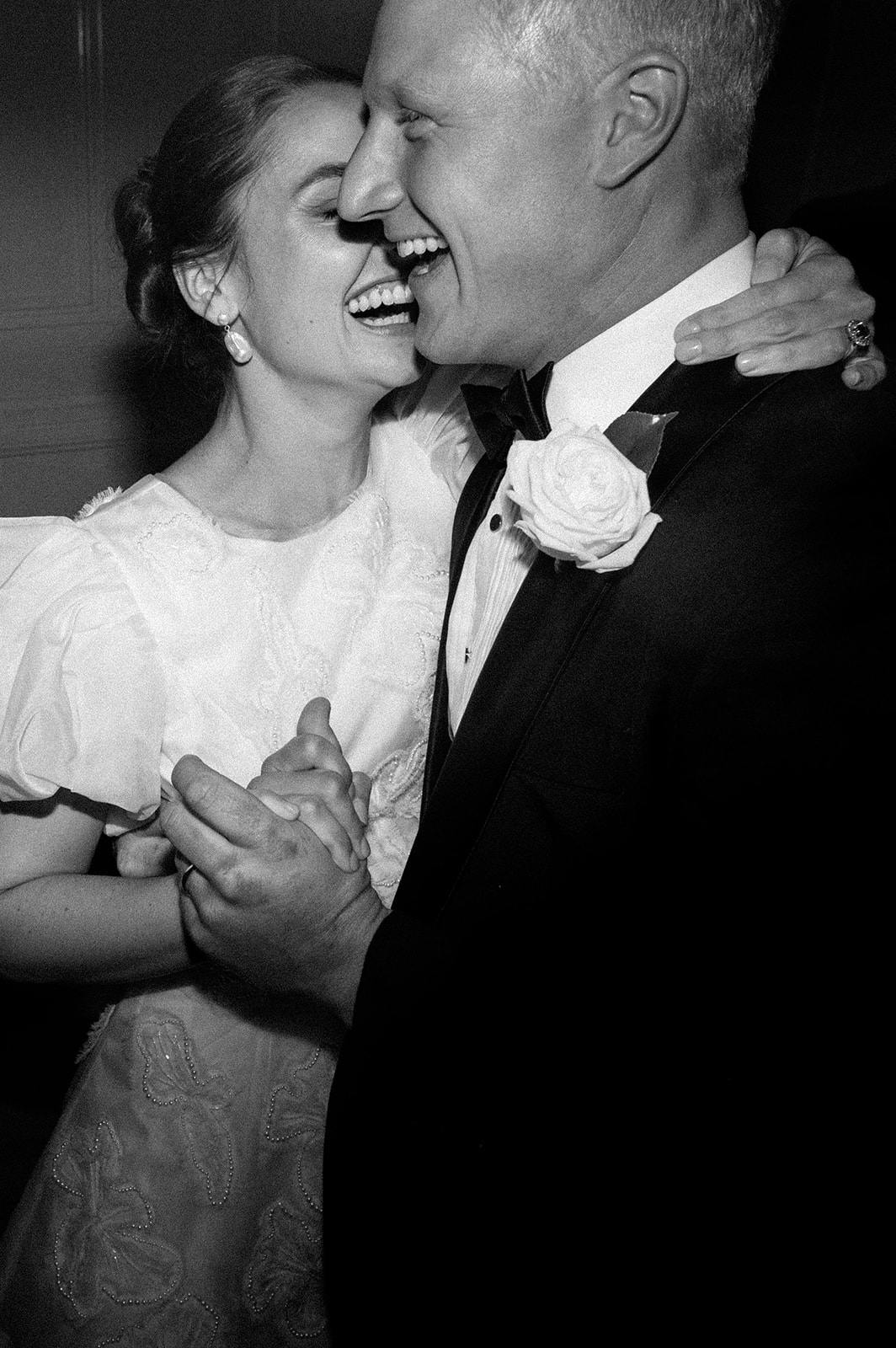 A black-and-white photo of a couple dancing and laughing joyfully. The woman wears a white dress with puffed sleeves, and the man is in a black suit with a bow tie and a rose on the lapel. They are holding hands, with their faces close together, sharing a happy moment.