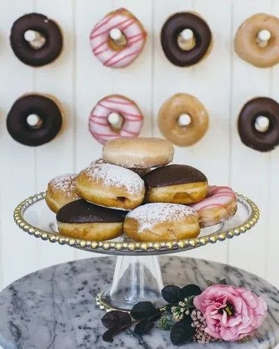 A decorative display of donuts. On a glass cake stand, an assortment of donuts with chocolate, powdered sugar, and pink glaze are arranged. In the background, there are various glazed and chocolate-topped donuts hanging on a white wall. A pink flower lies on the table.