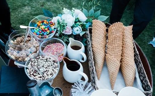 A table set up for an ice cream sundae bar with waffle cones, chocolate and rainbow sprinkles, candy pieces, and other toppings in bowls. A bouquet of flowers is partially visible in the background.