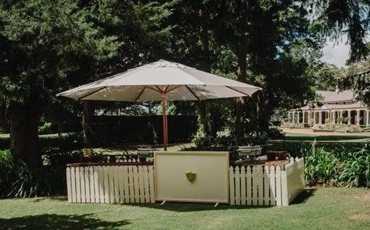 A quaint outdoor setup with a large white umbrella shading a rustic bar enclosed by a white picket fence. The bar is surrounded by lush greenery and tall trees, with a traditional-style house visible in the background.