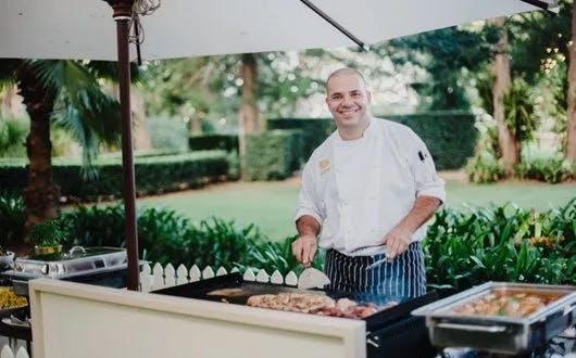 A chef in a white uniform and striped apron is smiling while cooking on a large outdoor grill. He is under a white umbrella and is surrounded by lush greenery and a garden setting. Various dishes can be seen on the grill and on serving trays nearby.