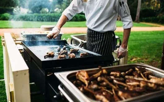 A chef wearing a white uniform and striped apron is grilling food on an outdoor flat-top grill. The chef is using tongs to flip the food. In the foreground, there is a chafing dish filled with cooked meat. The background features green grass and trees.