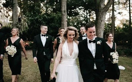 A bride in a white dress and a groom in a black tuxedo walk outdoors surrounded by their wedding party. The bridesmaids wear black dresses and hold white bouquets, while the groomsmen wear black tuxedos. The background features trees and greenery.