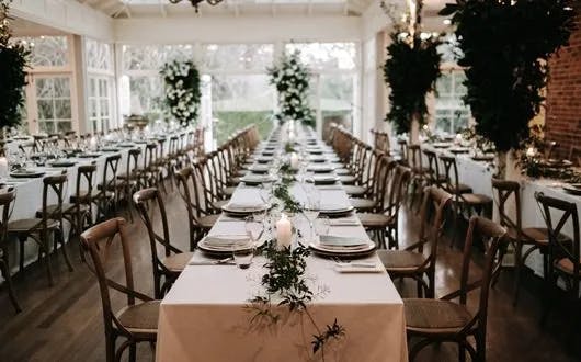 A beautifully decorated dining area with long tables covered in white tablecloths, set with plates and cutlery. Greenery and candles adorn the tables. Wooden chairs are neatly arranged along the sides. Large windows and plants create a bright and elegant ambiance.