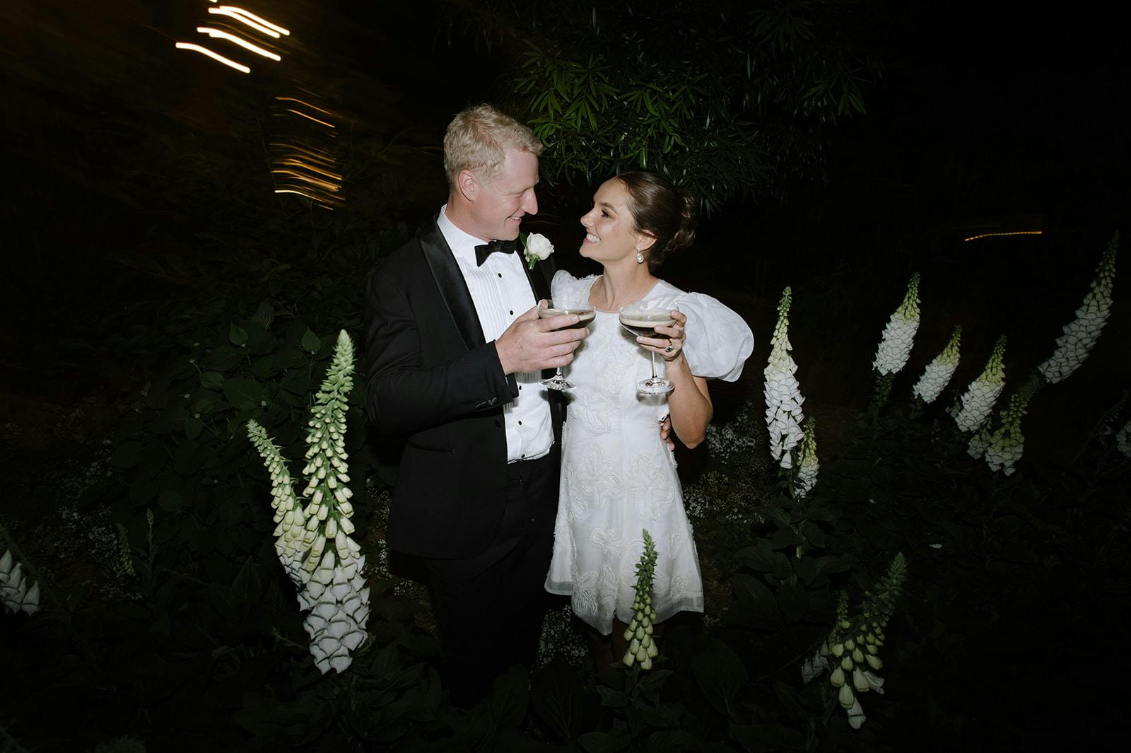 A couple, dressed elegantly in a black tuxedo and a short white dress, smile at each other while holding up champagne glasses. They stand amidst tall, white flowers at night, with blurred lights in the background adding a romantic glow to the scene.