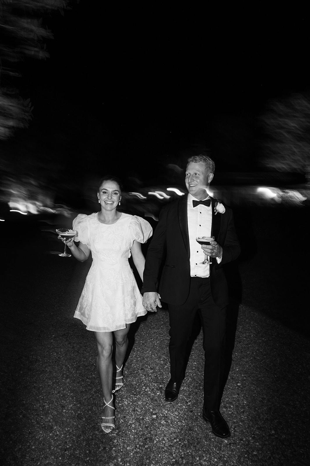a newly married couple walking through the grounds of gabbinbar at night holding drinks