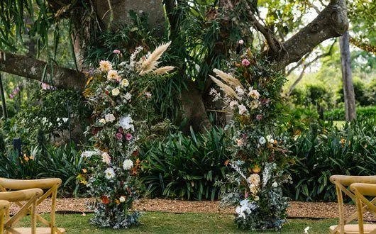 A lush outdoor wedding setup featuring two tall, decorative floral arrangements with a variety of colorful flowers and greenery. The arrangements frame a large tree in the background. Simple wooden chairs are positioned on either side, creating an aisle.