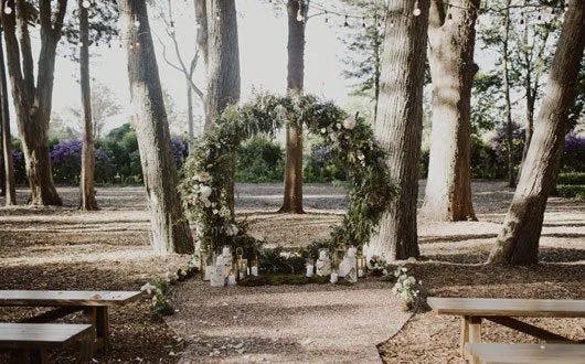 A wedding ceremony setup in a forest features a large, circular floral arch decorated with lush green foliage and white flowers. Wooden benches are arranged for seating, and the ground is covered in pine needles. Strings of lights are hung above.