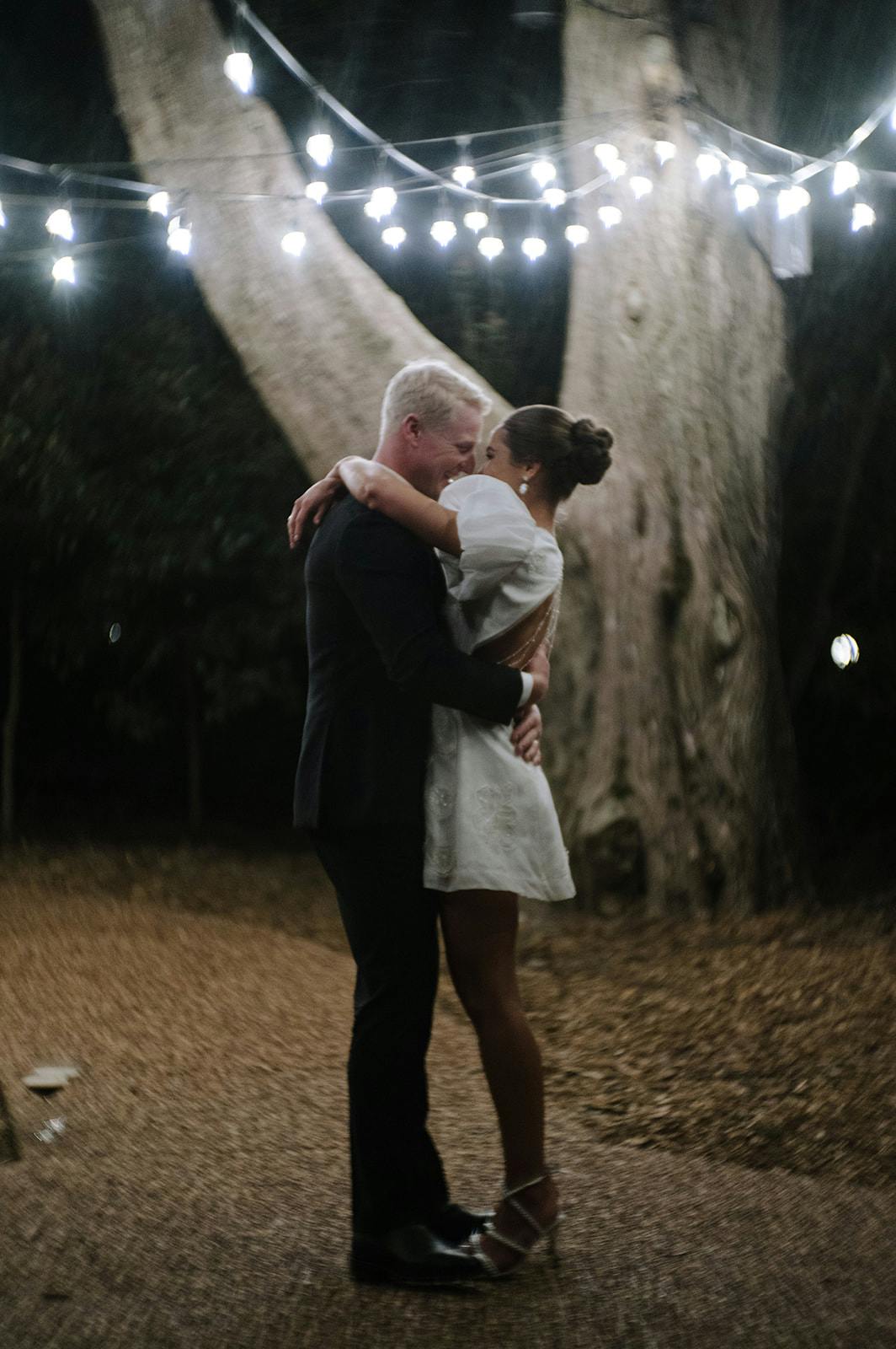 A couple is embracing under string lights at night. The woman is wearing a white dress and the man is in a dark suit. They are standing on a dirt path surrounded by trees, creating a romantic and intimate atmosphere.
