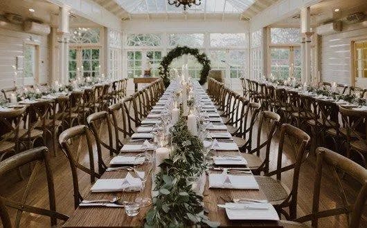 A long, wooden table set for a banquet is the focal point, positioned in the center of a bright, airy, glass-enclosed room. The table is decorated with white linens, elegant place settings, and a lush green garland. Rows of chairs line both sides of the table.