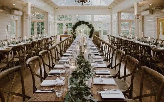 A long, wooden table set for a banquet is the focal point, positioned in the center of a bright, airy, glass-enclosed room. The table is decorated with white linens, elegant place settings, and a lush green garland. Rows of chairs line both sides of the table.