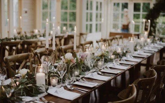 Elegant dining tables set for a special event. Tables are adorned with white candles, flower arrangements, and greenery, creating a romantic ambiance. The room is well-lit with natural light coming through large windows in the background.