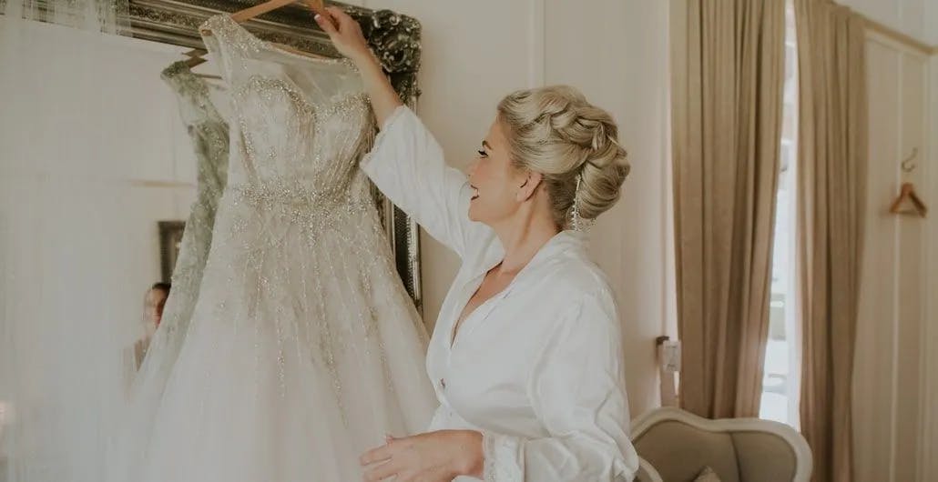A smiling bride dressed in a white robe reaches to hang her ornate, beaded wedding gown on a mirror in a softly lit room with beige curtains. The room has plush seating and creates an elegant, serene atmosphere suitable for wedding preparations.
