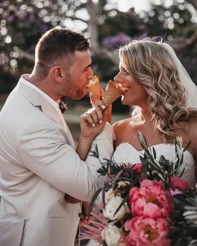 A bride and groom in wedding attire hold ice cream cones and playfully feed each other. They stand outdoors with trees in the background. The bride holds a colorful bouquet with pink and green flowers and smiles at the groom. Both appear joyful and relaxed.