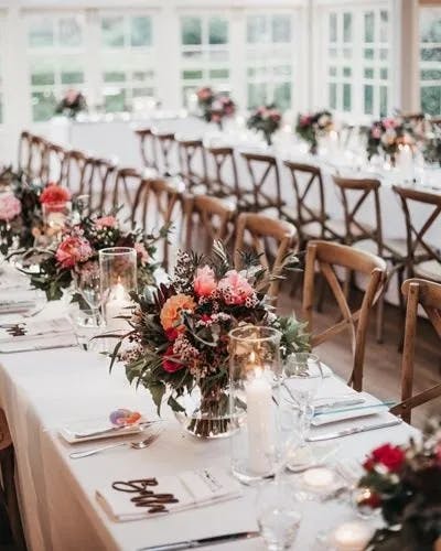 A beautifully decorated event venue with long dining tables adorned with floral centerpieces featuring pink and red flowers, candles, and elegant tableware. Wooden chairs are neatly arranged along the tables against a backdrop of large windows.