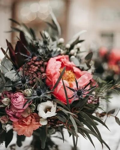 A vibrant floral bouquet featuring a mix of pink peonies, white lisianthus, and various greenery, including eucalyptus leaves and thistle stems. The background is softly blurred, drawing attention to the detailed textures and colors of the flowers.