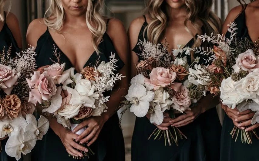 Four people in black dresses stand closely together, each holding a bouquet of flowers. The bouquets feature a mix of white, pink, and peach-colored blossoms, along with various greenery and dried elements. Their heads are not visible in the image.