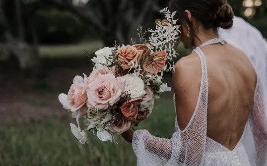A bride in a white, backless wedding dress is holding a large bouquet of pink and peach roses with greenery. The scene is set outdoors with blurred trees in the background.