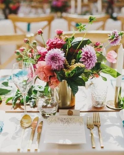 A beautifully set table with a floral centerpiece featuring pink and purple flowers in a gold vase. The table is set with wine glasses, water glasses, gold cutlery, and a menu card. The background has more tables with similar decorations.