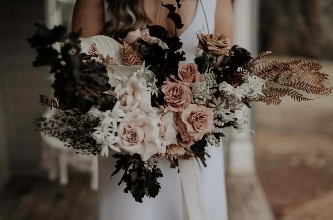 A woman in a white dress holds a large bouquet of flowers, featuring blush roses, white blooms, and dark leafy foliage, creating a rustic and elegant arrangement. The background is softly blurred.