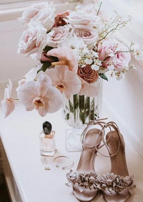 A delicate arrangement featuring a glass vase with pink and white roses, and orchids. Nearby are a pair of beige sandals adorned with floral embellishments, a bottle of perfume, a bracelet, and two pairs of earrings, all placed on a white surface.