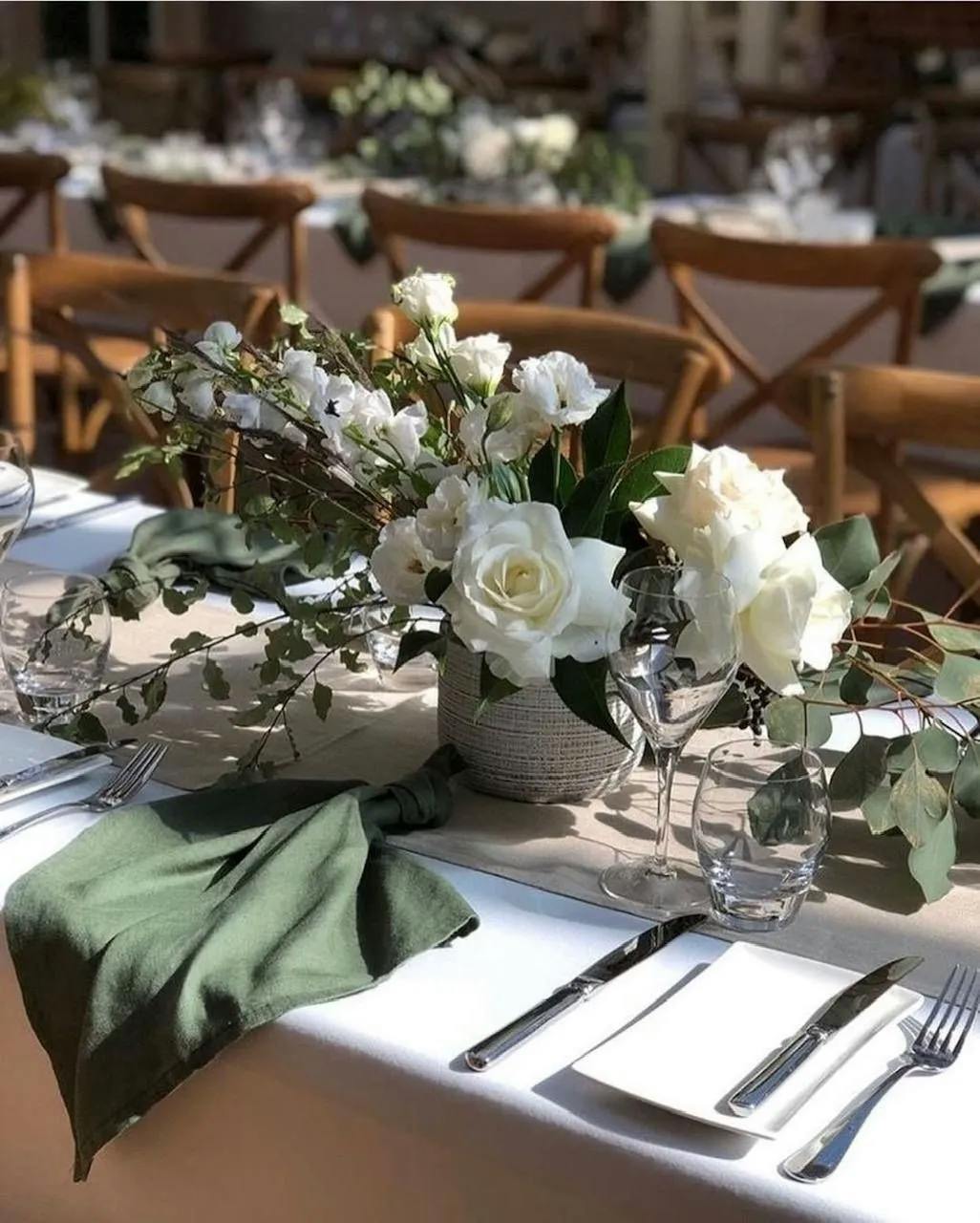 A close-up of an elegantly set dining table for an event. The table features a centerpiece with white roses and greenery. Each place setting includes silver cutlery, a wine glass, water glass, and a dark green napkin. Brown wooden chairs line the table.