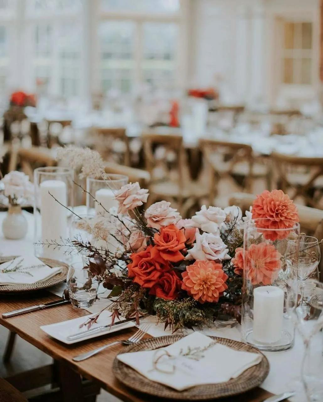 A beautifully set long dining table decorated with a centerpiece of red, peach, and white flowers. There are white candles in glass holders, woven placemats, and elegantly folded napkins arranged throughout. The background shows more similarly set tables.