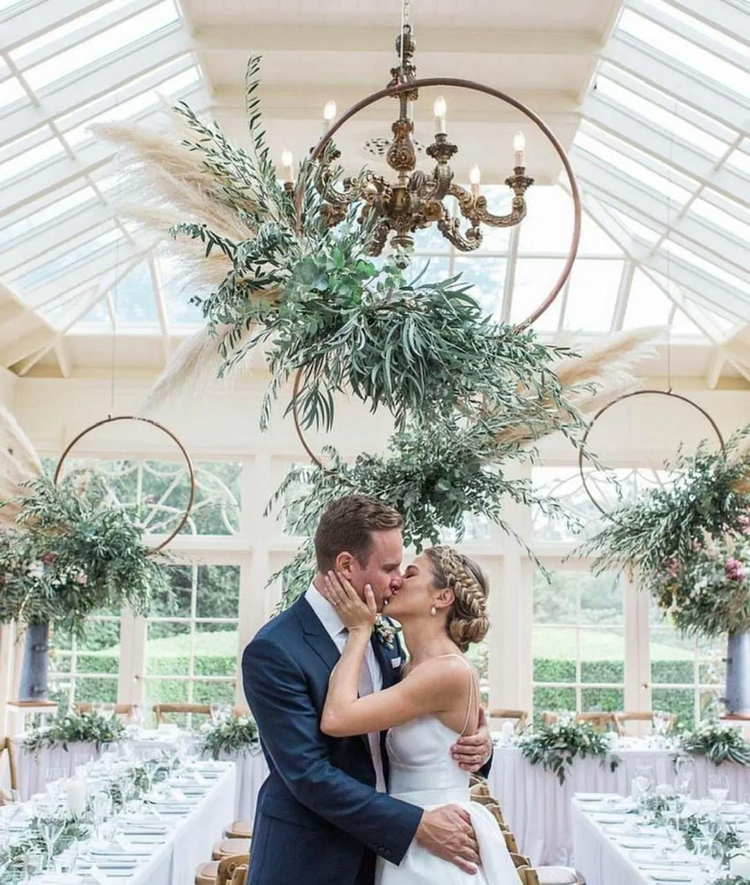 A bride and groom share a kiss under a chandelier adorned with greenery and flowers. They are in an elegantly decorated venue with large windows, circular floral arrangements, and long tables set for a wedding reception. The bride wears a white gown and the groom wears a blue suit.