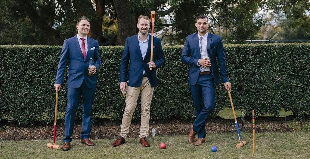 Three men in suits pose for a photo while holding croquet mallets. The man in the center stands with the mallet resting on the ground, while the other two hold drinks. They are outdoors with a neatly trimmed green hedge and trees in the background.