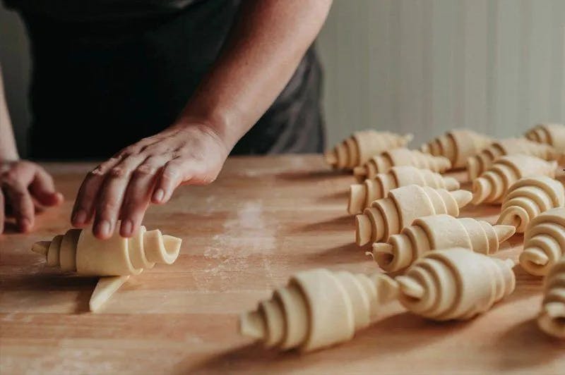 Person rolling croissant dough on a wooden surface, with several unbaked croissants arranged in the background. The hands are actively shaping one of the croissants, creating a sense of meticulous preparation in a cozy kitchen setting.
