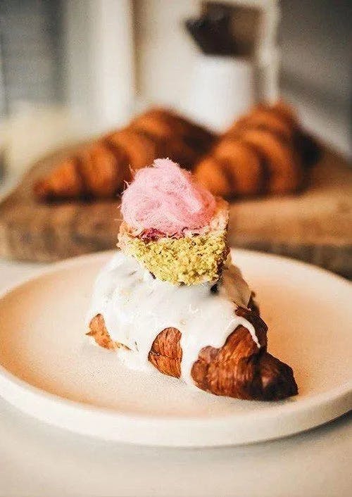 A decorated croissant sits on a white plate. The croissant is topped with white icing, a pistachio-encrusted pastry, and a dollop of pink cotton candy. In the blurred background, a cutting board with additional croissants is visible.