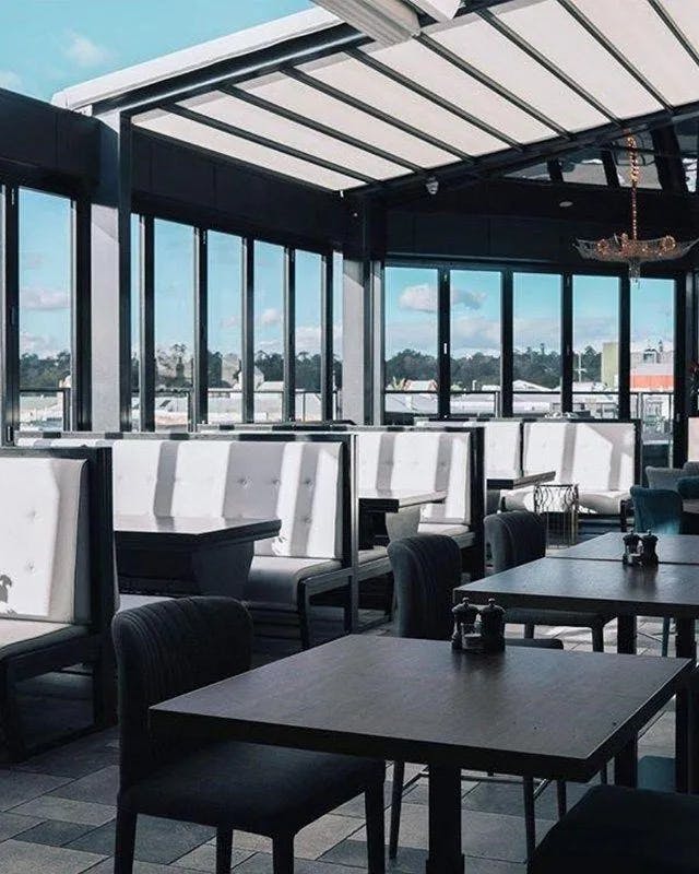 A modern indoor dining area featuring upholstered white booths along the windows and dark wooden tables with black chairs. Large windows and a glass ceiling allow natural light to flood the space, providing a clear view of a sunny, blue sky outside.
