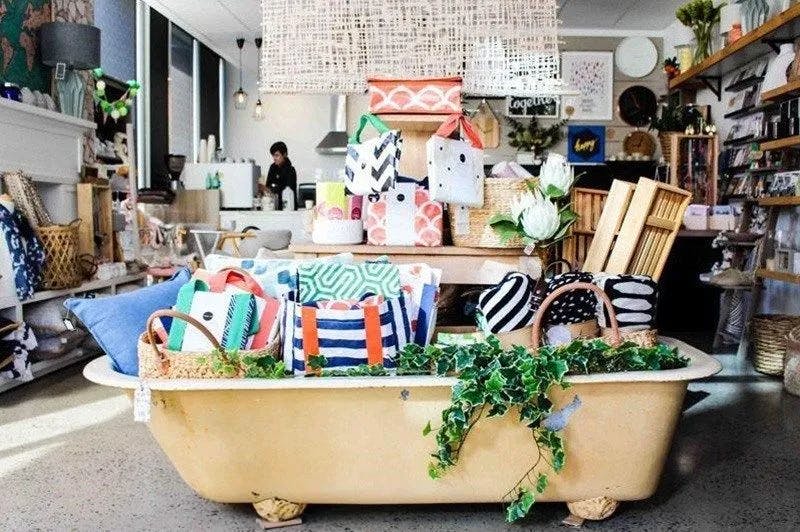 A cozy store with a variety of colorful handmade bags and decorative pillows displayed inside a vintage clawfoot bathtub. The bathtub is adorned with green ivy and surrounded by shelves filled with other eclectic items. A person is seen working in the background.