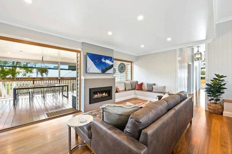 A spacious, modern living room featuring a dark brown leather couch, a built-in fireplace, and a wall-mounted TV. Adjacent is a large sliding door leading to an outdoor dining area with a table and chairs. The room has light wood floors and bright decor.