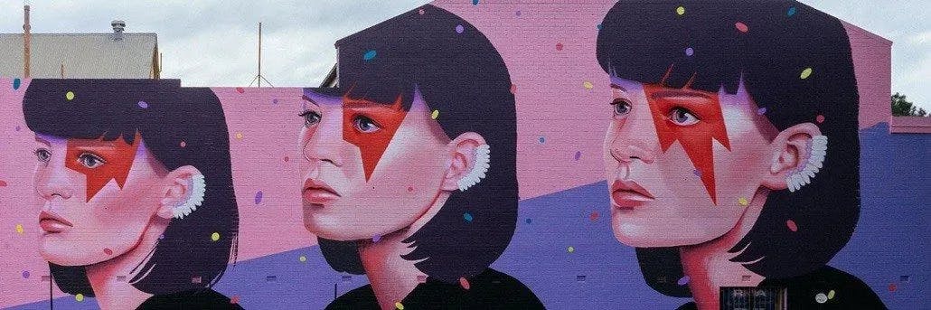 A mural featuring three side-by-side portraits of a woman with short black hair, red facial markings resembling lightning bolts, and white ear adornments. The background is a gradient of pink to purple, with scattered colorful confetti-like dots.