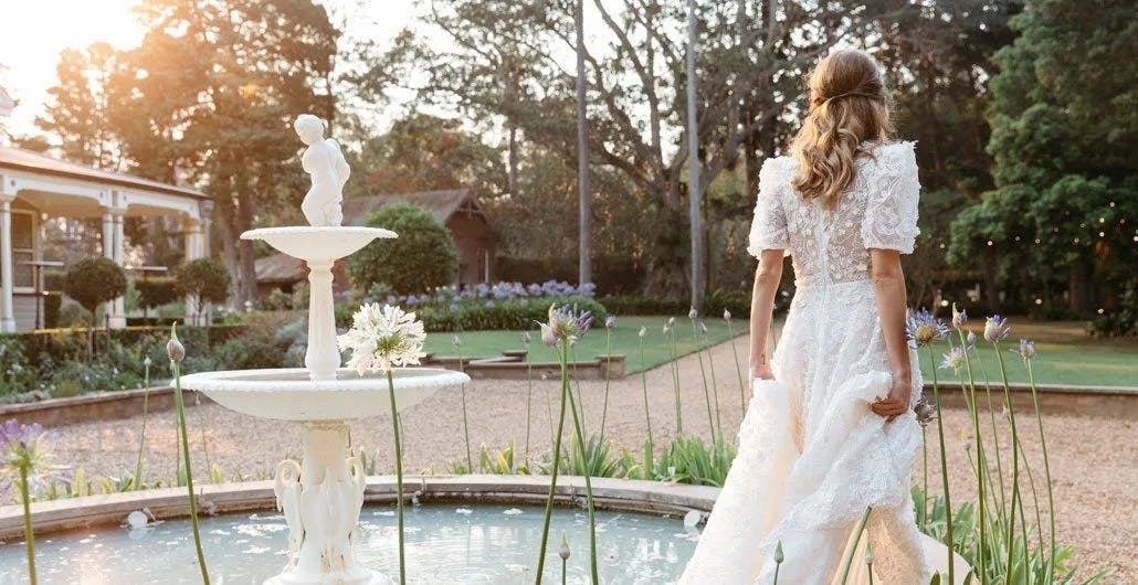 A bride in a white lace wedding dress stands by a picturesque fountain in a garden, holding up her gown slightly. The sun is setting, casting a warm glow over the scene, which includes greenery and a charming house in the background.