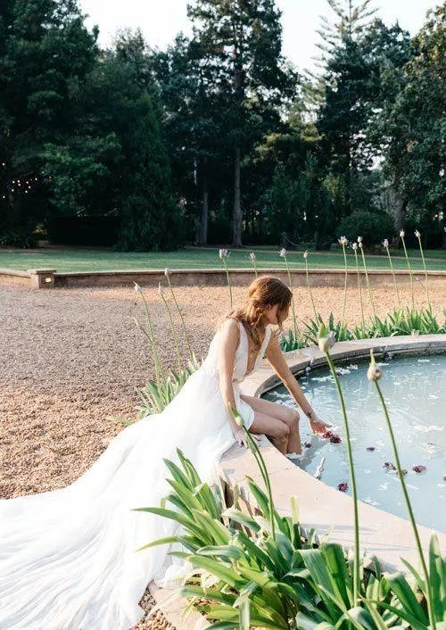 A woman in a flowing white dress sits at the edge of a circular fountain surrounded by greenery and trees. She dips her hand into the water, creating gentle ripples as flower petals float on the surface. The scene is serene and picturesque.