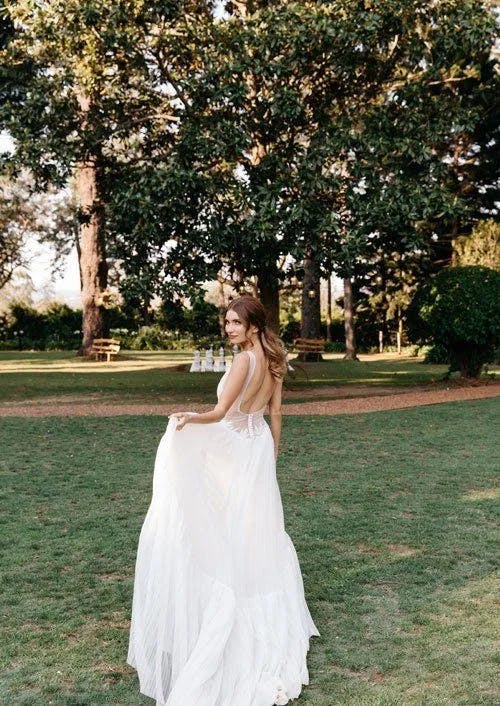 A bride in a flowing white wedding dress stands on a green lawn, looking back over her shoulder. She is surrounded by tall trees and greenery in a serene outdoor setting. The sun is shining, casting a soft light on the scene.