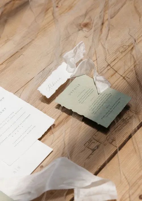 A close-up photo of scattered wedding invitations on a wooden table. The invitations are partially covered by a sheer, white fabric. One green invitation with the word "Details" is prominently displayed, alongside a small piece of paper with the handwritten name "Rebecca".