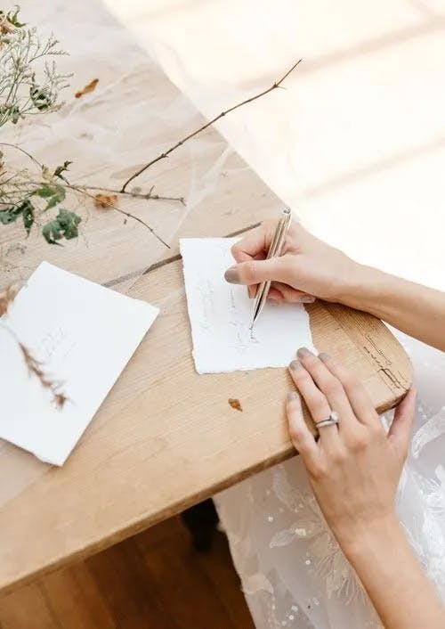 Close-up of a person's hands writing on a piece of paper with a pen. The person is also wearing a ring on their left hand. The table has dried flowers and another piece of paper. Soft, natural light illuminates the scene.
