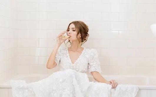 A woman in a white wedding dress sits inside a bathtub against a white tiled wall, sipping from a glass. She has her hair styled in an updo and is looking content and relaxed.