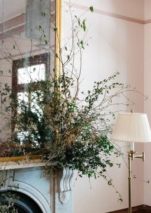 A large, ornate mirror with a gold frame hangs above a white marble fireplace. An arrangement of green and dried branches extends from the mantelpiece. A white floor lamp with a gold base stands to the right, illuminating the room with soft light.