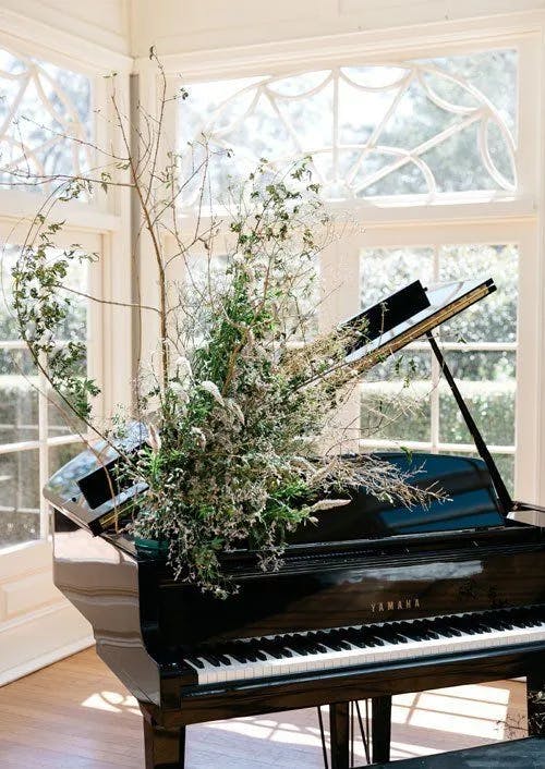 A grand piano with the lid open is positioned in a sunlit room with large windows. The piano is adorned with a lush arrangement of green foliage and white flowers, which extends from the interior. The scene exudes an elegant and serene ambiance.