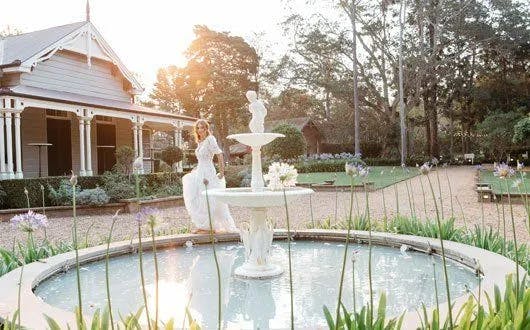 A woman in a white wedding dress stands beside a two-tiered fountain in a garden. The sun is setting, casting a golden light over the scenic background, which includes a charming wooden house with a veranda and tall trees around the garden.