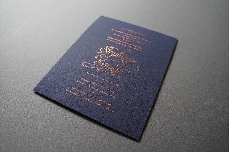 A close-up of a navy blue wedding invitation with elegant gold script. The names "Stephane & Ernesto" are prominently featured in the center, with additional wedding details written above and below the names. The invitation rests on a smooth, grey surface.