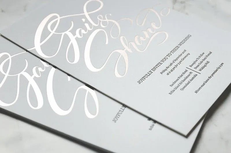 A close-up of a wedding invitation with elegant silver cursive text. The top portion features stylized names in large, flowing script, while the bottom portion includes details about the date, time, venue, and additional wedding information in smaller, simple print.